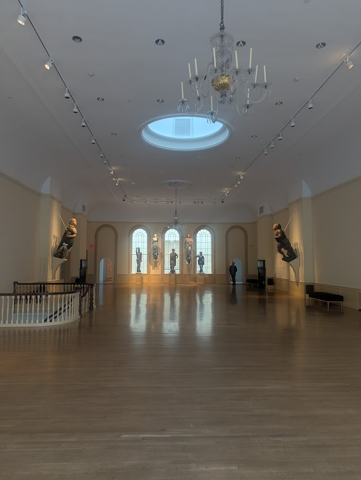 A very large hall in the museum with cool statues on the wall. They were playing cool music in the room that echoed.